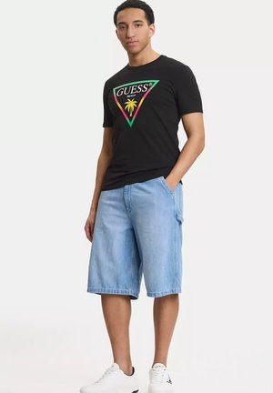 Hombre con camiseta negra con logotipo de triángulo verde y rosa y palmera, pantalones cortos de mezclilla azul claro y zapatillas blancas, de pie de manera relajada.