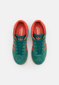 Green suede sneakers with orange accents, featuring three stripes, round toe, lace-up front, and a textured rubber sole.