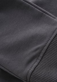 Donkergrijze stof met een close-up van de gestikte naad en ribbelmanchetten op de mouw van een gebreid kledingstuk.