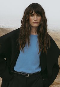 Melna pārmērīga izmēra mēteli virs gaiši zila džempera, papildināta ar melniem augstajiem biksēm un plānu melnu jostu, ar teksturētām viļņveida matiem.