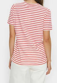 Mujer con una camisa blanca de mangas cortas con rayas horizontales rojas y pantalones blancos, de pie de espaldas a la cámara.