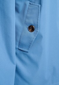 Světle modrá látka s hladkou texturou, zdobená našitou kapsou a tmavě hnědým knoflíkem se čtyřmi dírkami, zdůrazňující funkční designové prvky.