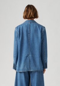 Denim blejzer v srednje modri barvi z prilagojenim krojem, enim zadnjim ventom in gumbnimi manšetami. Ima izrezan ovratnik in subtilno teksturo.