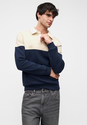 Jeune homme portant un polo à manches longues crème et marine ainsi qu'un jean gris, posant avec une main près du menton et regardant sur le côté sur un fond blanc.