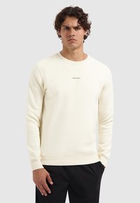 Man met krullend haar in een crèmekleurige sweatshirt met de tekst "PURE PATH" en zwarte broek, staande tegen een effen lichtgrijze achtergrond.