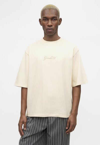 Άνδρας με κοντά ξανθά μαλλιά φοράει φαρδιά εκρού μπλούζα με το κείμενο "GmbH" και μαύρο-λευκό παντελόνι με κάθετες ρίγες.