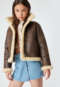 Braune Kunstlederjacke mit beigem Lammfellkragen und -manschetten. Reißverschluss, cropped Design, kombiniert mit einem hellblauen Jeansrock.