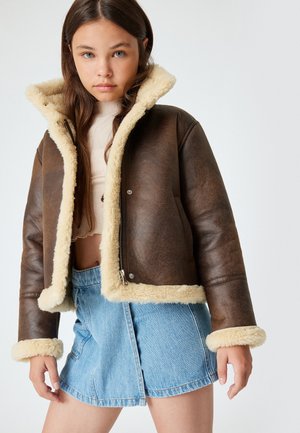 Giacca in finta pelle marrone con colletto e polsini in shearling crema. Chiusura con zip, design cropped, abbinata a una gonna di jeans azzurri chiara.