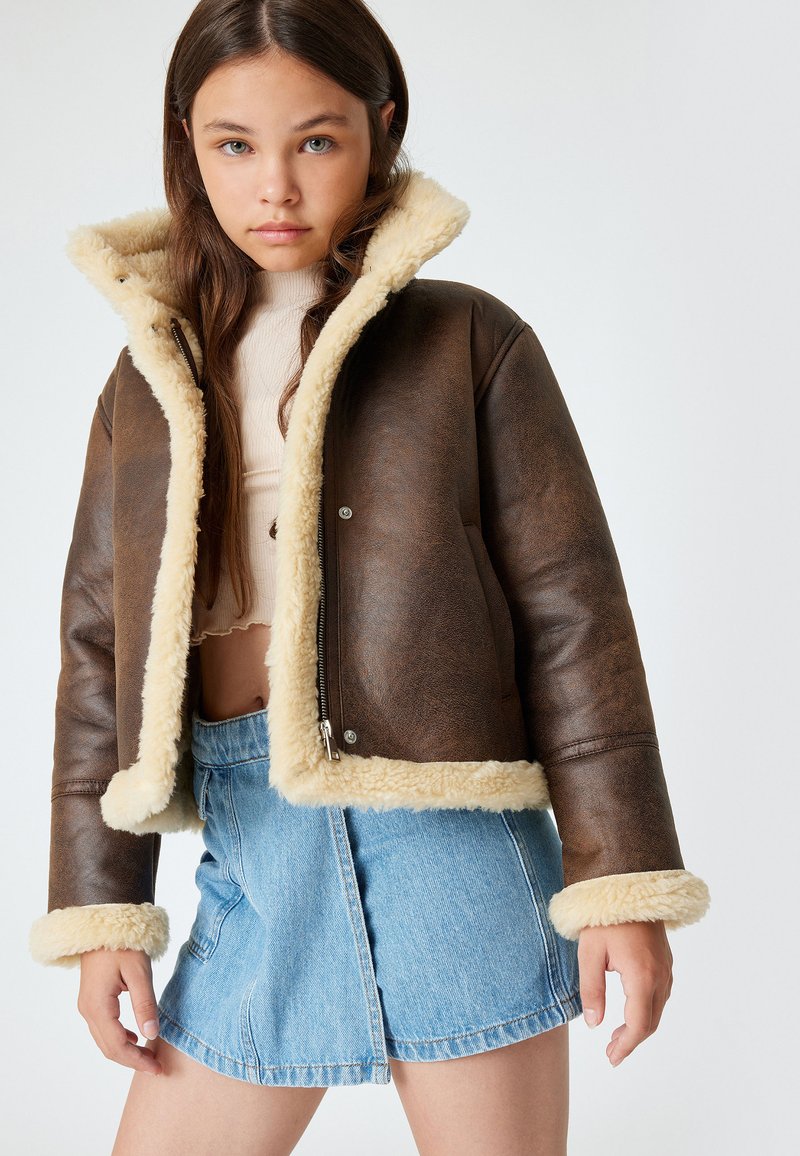 Braune Kunstlederjacke mit beigem Lammfellkragen und -manschetten. Reißverschluss, cropped Design, kombiniert mit einem hellblauen Jeansrock.