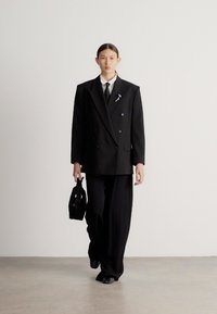 Μαύρο διπλόμπουτο σακάκι συνδυασμένο με φαρδιά παντελόνια, λευκό πουκάμισο και λεπτή μαύρη τσάντα. Διακριτική καρφίτσα στο πέτο του σακακιού.