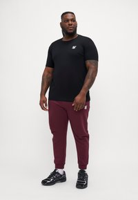 T-shirt scurt negru cu un logo pe piept, asortat cu pantaloni sport vișinii. Modelul poartă adidași negri.