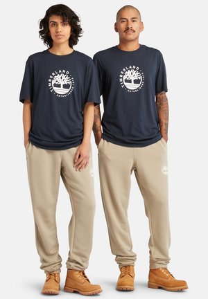 Marineblauwe katoenen T-shirts met een wit logo, beige joggingbroeken en bruine laarzen; ontspannen pasvorm met zichtbare stiksels en geborduurde accenten.
