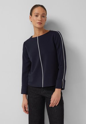 Kobieta ubrana w granatową bluzkę z długim rękawem z białymi przeszyciami na środku i na rękawach, w czarnych spodniach, stoi na tle szarej ściany.