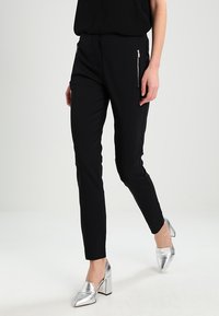 Pantalons noirs ajustés avec poches zippées, coupe slim et texture lisse. Associés à des chaussures à talons hauts argentées et brillantes.