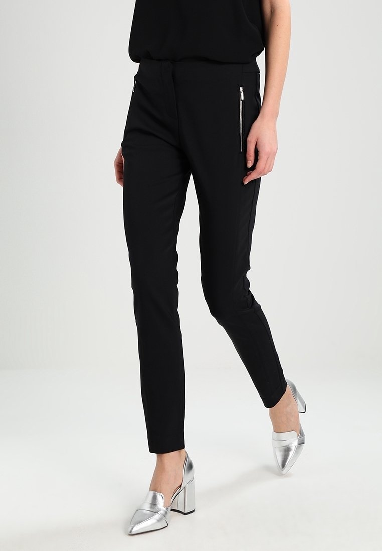 Pantalons noirs ajustés avec poches zippées, coupe slim et texture lisse. Associés à des chaussures à talons hauts argentées et brillantes.