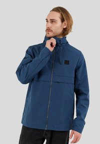 Chaqueta impermeable de color azul marino con cremallera frontal, capucha ajustable y bolsillo horizontal en el pecho con un parche de logo negro. Textura suave.