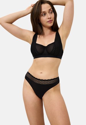 Femme portant un soutien-gorge en dentelle noire et un slip assorti, posant avec les bras levés contre un fond clair uni.