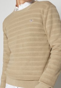 Beige gestrickter Pullover mit horizontalen Streifen, Rundhalsausschnitt und gerippten Bündchen. Mit kleinem Logo auf der Brust bestickt.