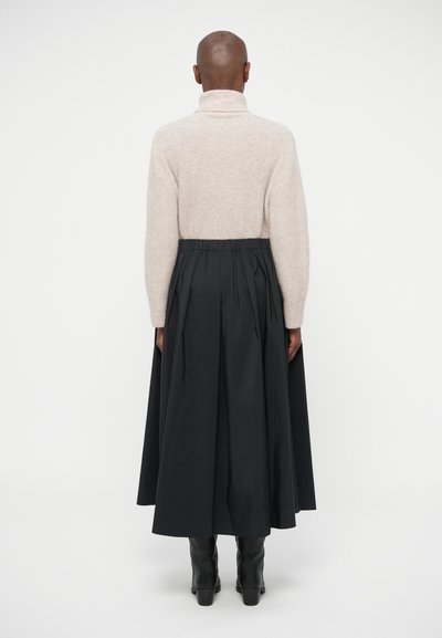 Lys beige strikket sweater med høj krave, parret med en lang sort plisseret nederdel. Model iført sorte ankelstøvler, set fra bagsiden.