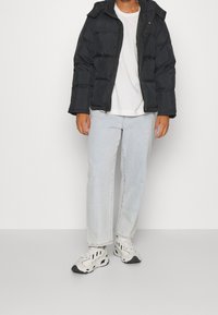 Doudoune noire avec capuche, dotée d'une fermeture éclair à l'avant et d'une texture matelassée. Portée avec un jean en denim clair et des baskets blanches.