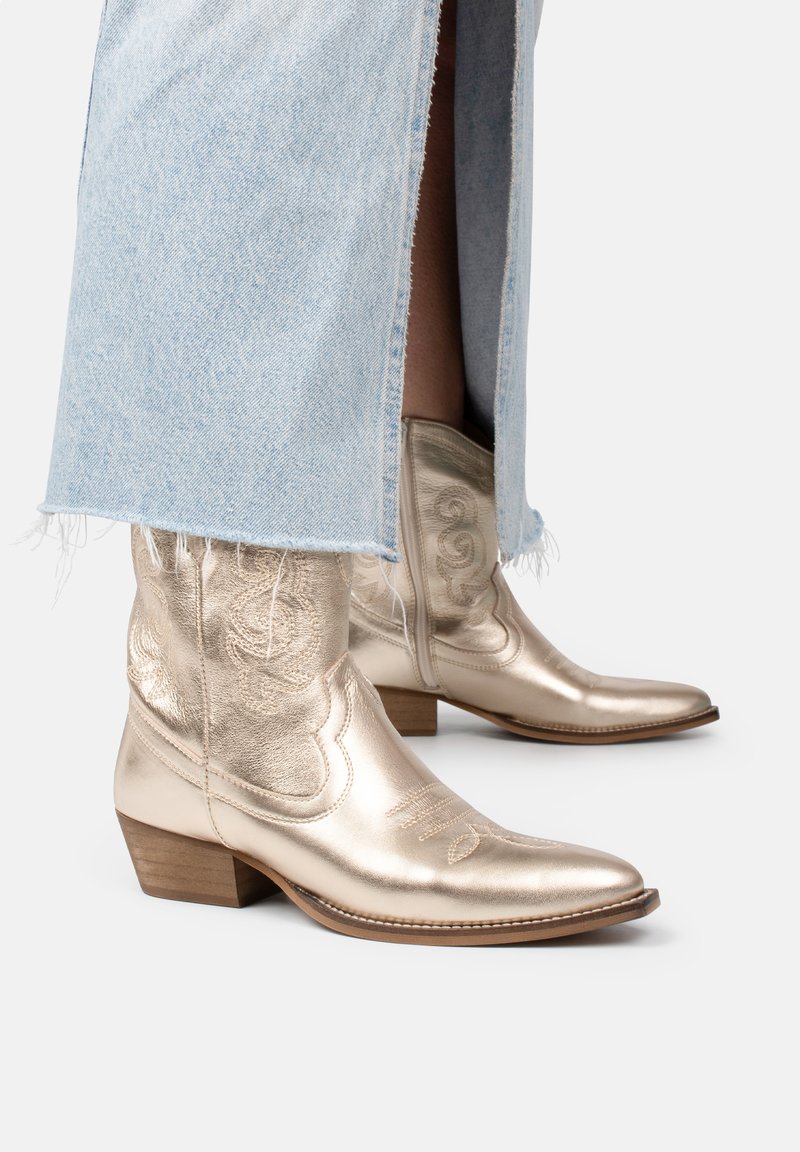 Botas de vaquero metálicas doradas con punta afilada y tacón bajo apilado, que cuentan con costuras decorativas y un acabado texturizado.