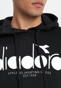 Sudadera negra de algodón acanalado, con un gran logotipo gráfico en blanco y texto. Incluye capucha con cordón y tejido texturizado.