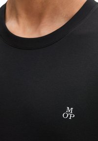 Κοντινό πλάνο μαύρου μπλουζάκι με στρογγυλή λαιμόκοψη και μικρό λευκό λογότυπο "M O'P" στη αριστερή πλευρά του στήθους, που φοριέται από άτομο.