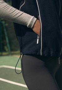 Tumesinine fliisvest, millel on hõbedane tõmblukk, valged torustikud ja küljetaskud. Kompressioonmustad retuusid reguleeritava nööriga ääres.