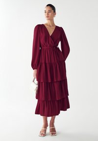 Burgundowa sukienka z falbanami, długimi bufiastymi rękawami, głębokim dekoltem w kształcie litery V i paskiem w talii. Połączona z białymi sandałami na paski i małą białą torebką.