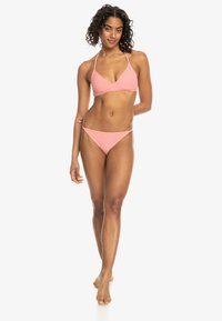 Roze bikini set met een gestructureerde driehoekstop en een lage broek, met dunne bandjes en minimale bedekking, op een model tegen een witte achtergrond.