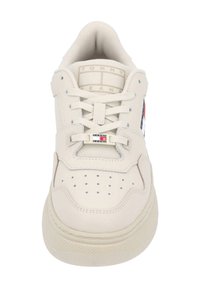 Béžová teniska s perforovanou špičkou, polstrovaným límcem a plochými tkaničkami. Má logo značky u jazyka a kontrastní detaily na bocích.