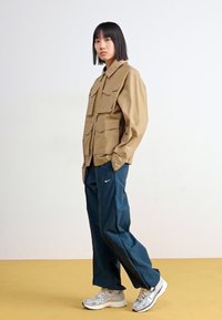 Ταν αδιάβροχο μπουφάν με μεγάλες μπροστινές τσέπες, συνδυασμένο με ναυτικές φόρμες και αθλητικά παπούτσια σε ασημί χρώμα. Ουδέτερη φόντο για αντίθεση.