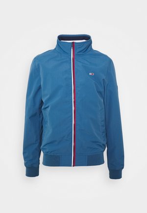 Niebieska kurtka na suwak z wysokim kołnierzem, ściągaczami na mankietach i dolnym brzegu, z małym prostokątnym logo na piersi i trójkolorowym suwakiem.