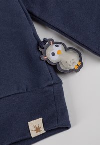 Felpa blu navy con tessuto testurizzato, caratterizzata da un'etichetta crema cucita e un tag a forma di gufo colorato attaccato.