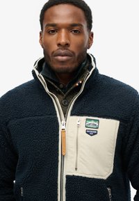 Giacca in pile blu navy con pannello crema, chiusura con cerniera e dettaglio tasche. Presenta toppe e una superficie testurizzata per l'isolamento termico.