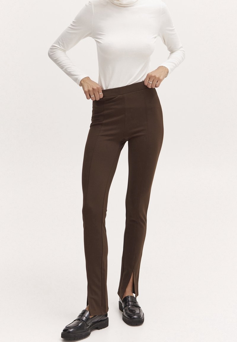 Personne portant un pantalon marron slim avec des fentes à l'avant des jambes, un haut blanc à manches longues et des mocassins noirs, debout devant un fond uni.
