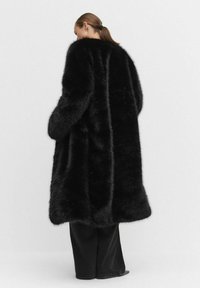 Svart konstpälsjacka med lös passform, långa ärmar och en mjuk, texturerad yta, når till mitten av låren, kombinerad med svarta byxor.