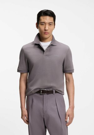 Mężczyzna w szarej koszulce polo włożonej do pasujących plisowanych spodni z czarnym paskiem, stojący na białym tle.