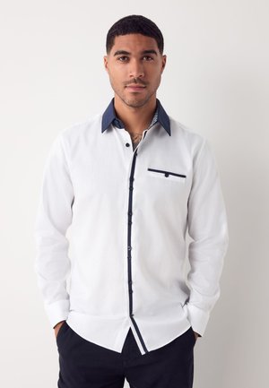 Valge pika varrukaga särk tekstuurse mustriga, kontrastse mereväe kraega ja nööbikonksuga ning väikese rinnanihiga.