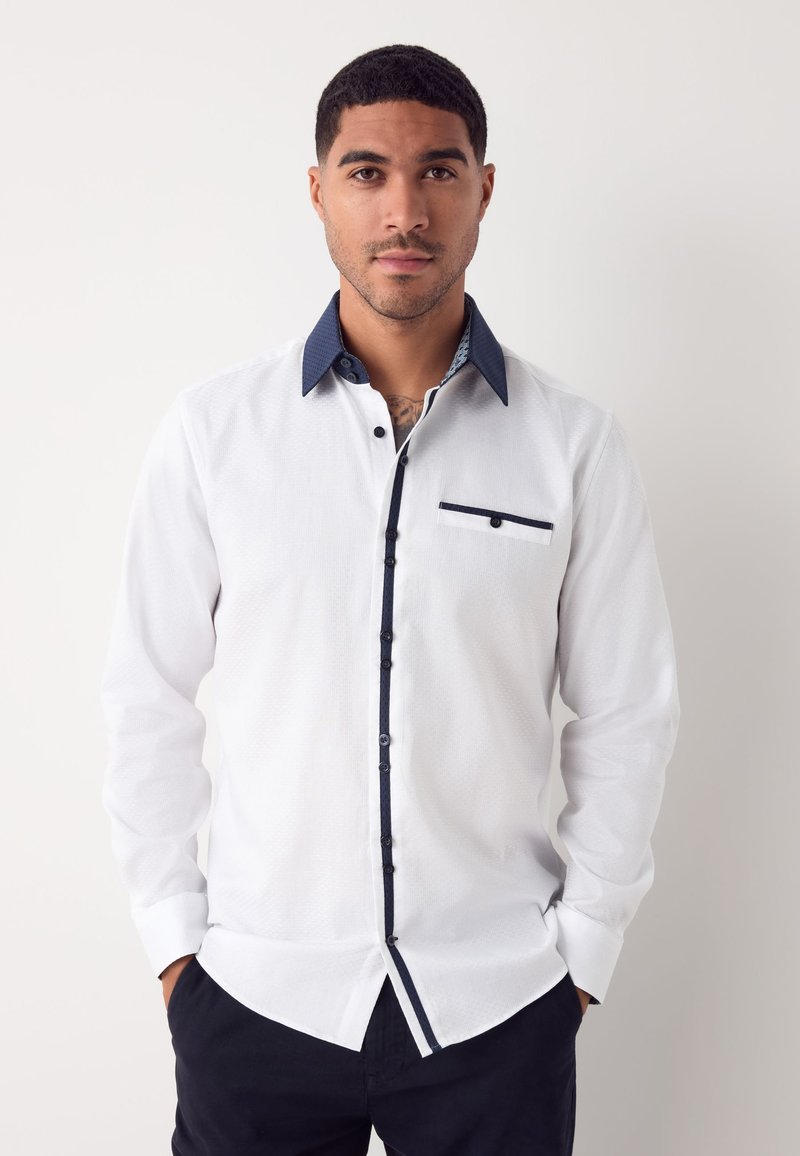 Camicia bianca a maniche lunghe con motivo testurizzato, colletto e patta dei bottoni a contrasto in navy, e una piccola tasca sul petto.
