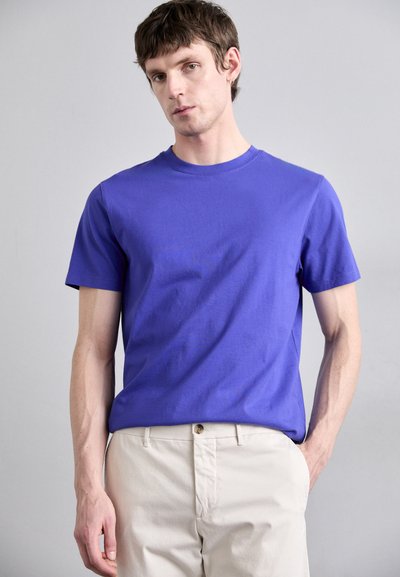 T-shirt violet à manches courtes en tissu lisse, associé à un pantalon plat de couleur claire. Design simple à col rond, sans motifs.