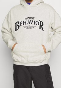 Lichtgrijze hoodie met zwarte bedrukte tekst "WORST BEHAVIOR" en een basketbalafbeelding. Hij heeft een kangoeroezak en ribbelboorden.