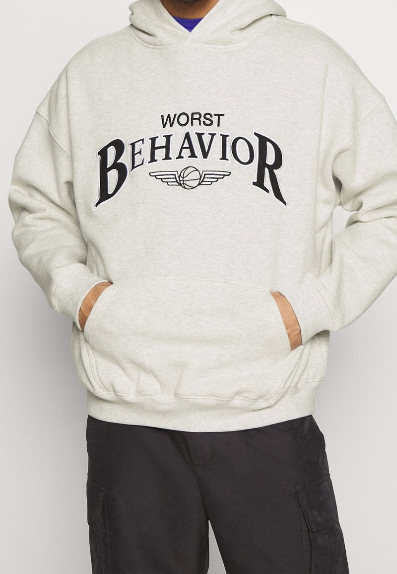 Lichtgrijze hoodie met zwarte bedrukte tekst "WORST BEHAVIOR" en een basketbalafbeelding. Hij heeft een kangoeroezak en ribbelboorden.