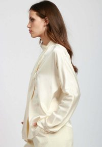 Blusa in satin colore crema con colletto alto, maniche lunghe e un leggero lucido. Presenta una chiusura frontale con un dettaglio di nappina decorativa.