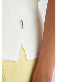 Witte katoenen T-shirt met zijsplit, met een O'Neill-label. Draagt over een getextureerd geel kledingstuk, dat de kleurcontrast en gelaagdheid benadrukt.