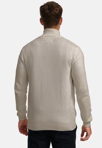 Man draagt licht beige gebreide trui met chevronpatroon, staat met zijn rug naar de kijker gekeerd tegen een effen achtergrond.