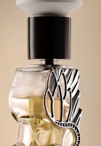 Bottiglia di profumo in vetro trasparente con design in foglie d'argento e tappo nero, contenente un liquido ambrato chiaro.