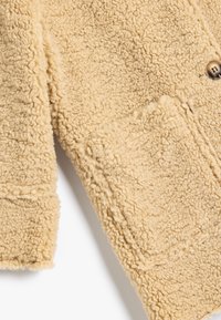 Beige faux shearling jakke med en tekstureret overflade, en enkelt knaplukning og en synlig forlommes. Blødt, varmt materiale.