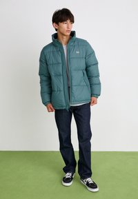 Zaļš pūkains jaka ar augstu apkakli, quilted dizainu, priekšējo rāvējslēdzēju un sānu kabatām, valkāts virs pelēkas krekla un tumšiem džinsiem.