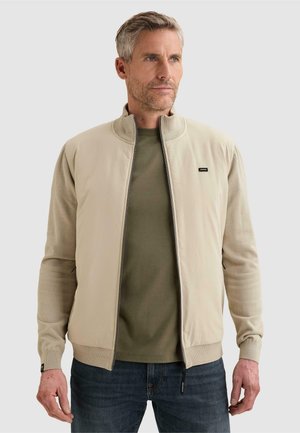 Mann trägt beige Zip-up-Jacke über olivgrünem Hemd und blauen Jeans vor schlicht hellem Hintergrund, schaut zur Seite.